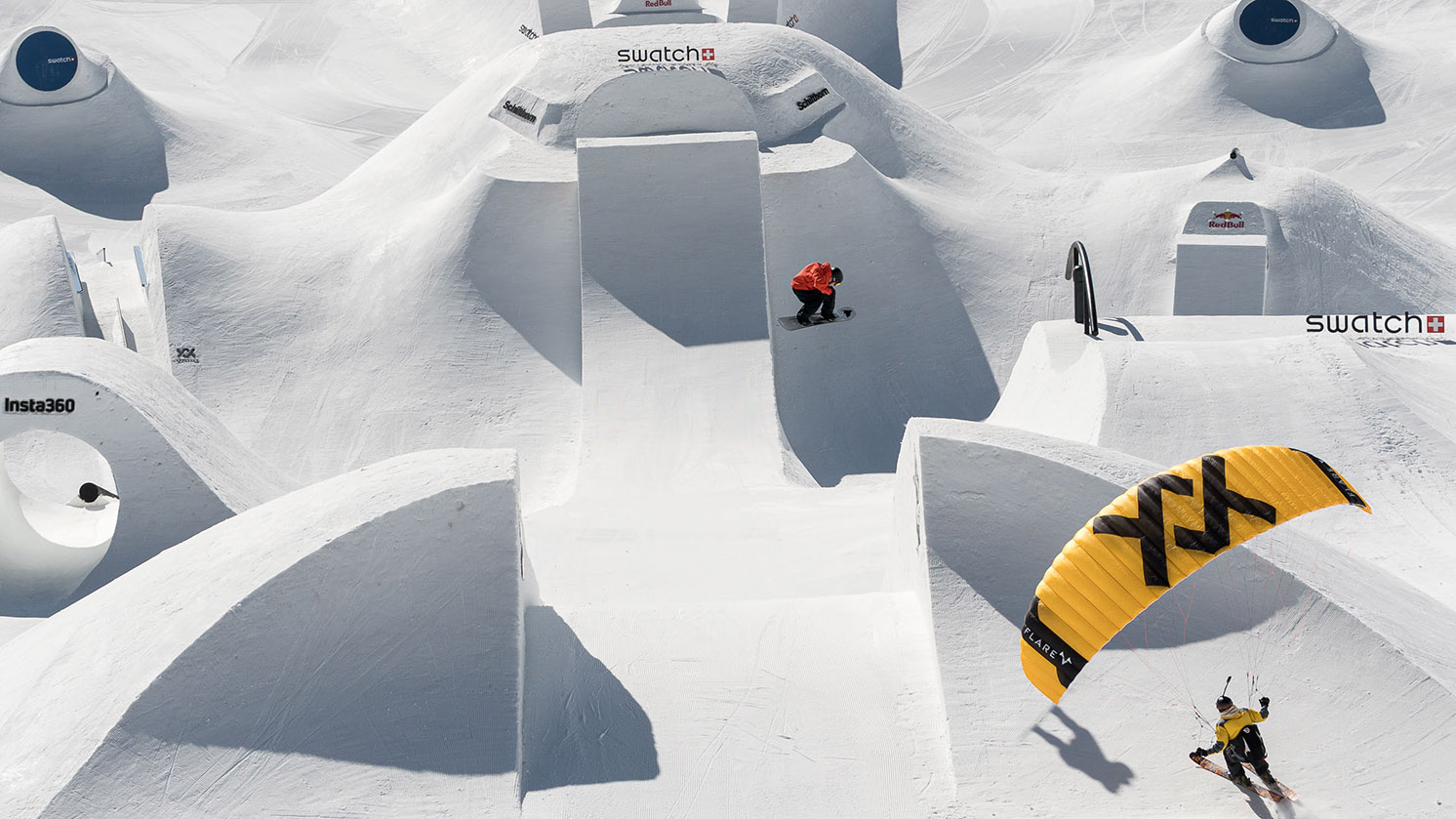 A skier at Swatch Nines 2025 gets big air while a paraglider flies in from the side.